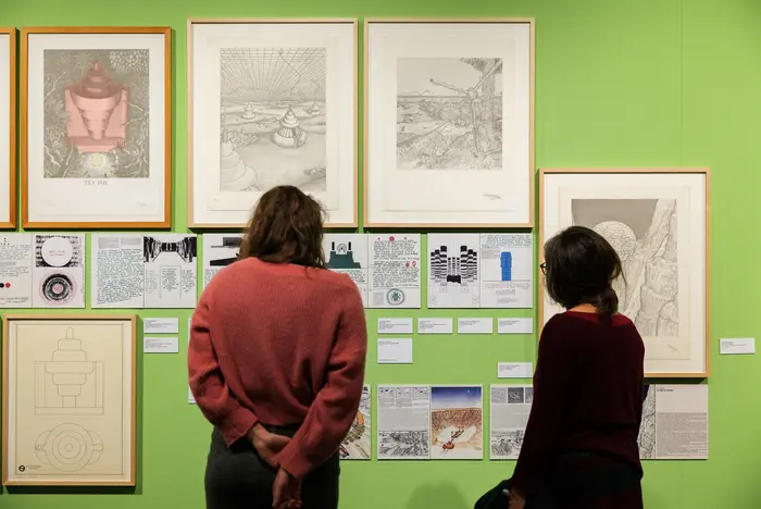 Zwei Besucher vor einer grünen Wand mit gerahmten Kunstwerken.