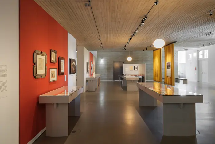 Ein Ausstellungsraum mit grauen Schaukästen und einer orangefarbenen Wand.