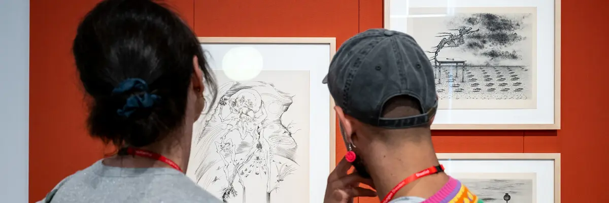 Zwei Besucher betrachten gerahmte Zeichnungen an einer orangefarbenen Wand.