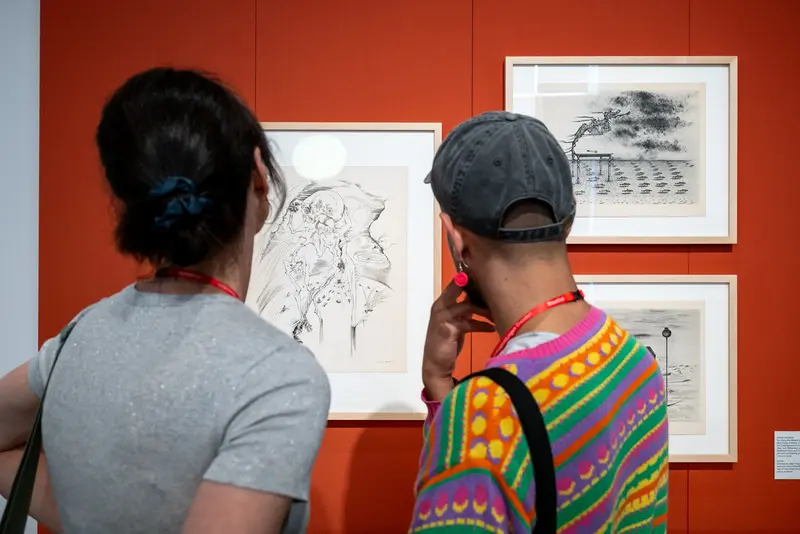 Zwei Besucher betrachten gerahmte Zeichnungen an einer orangefarbenen Wand.