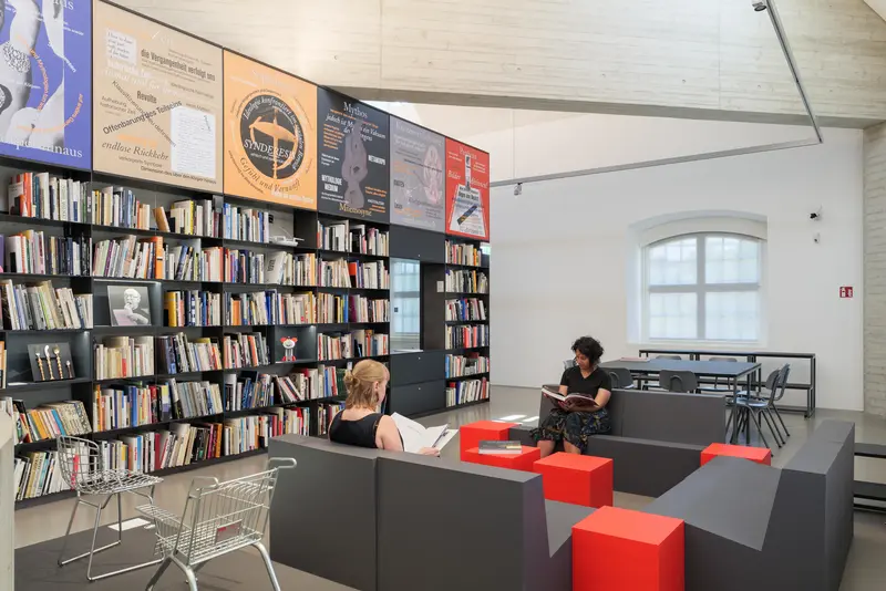 Zwei Personen sitzen in einem Raum mit Bücherregalen und Sofas.